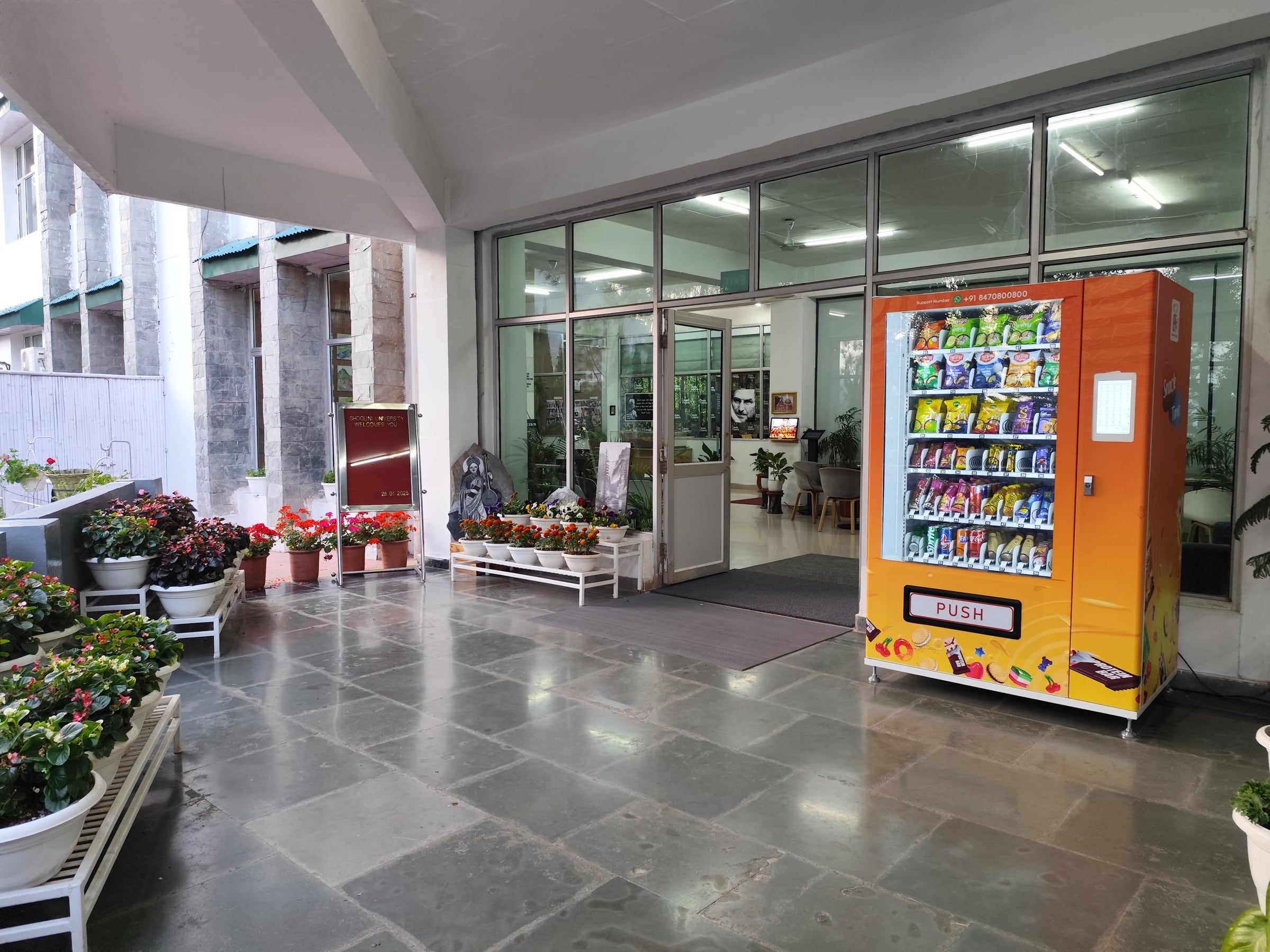 An orange Wendor vending machine at a university building entrance in daylight, with flower pots along the walkway