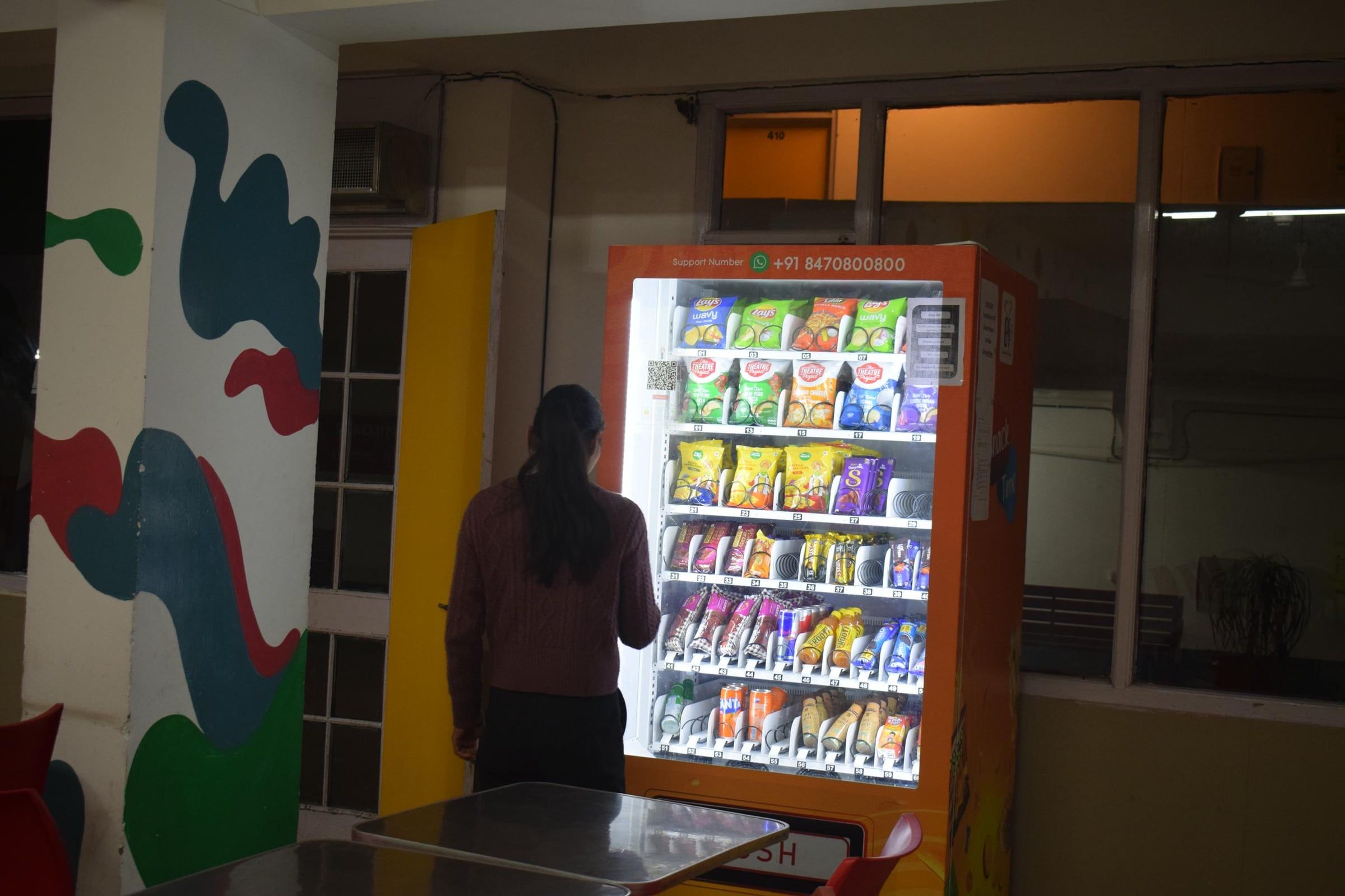 A student choosing snacks at a lit orange Wendor machine in a college canteen at dusk